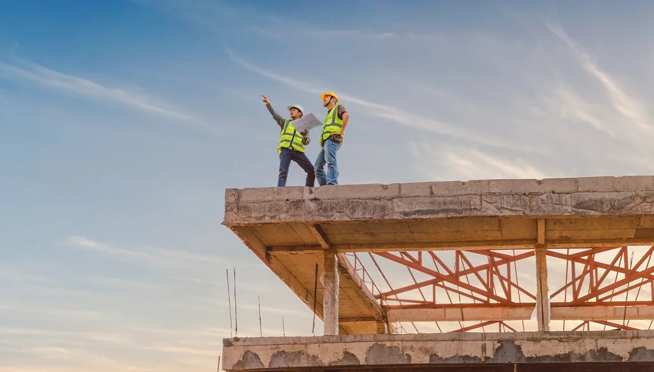 Twee bouwvakkers met veiligheidshesjes en helmen staan op de rand van een betonnen constructie in aanbouw. Eén houdt een bouwtekening vast terwijl de ander in de verte wijst. De lucht is helder met enkele sluierwolken.