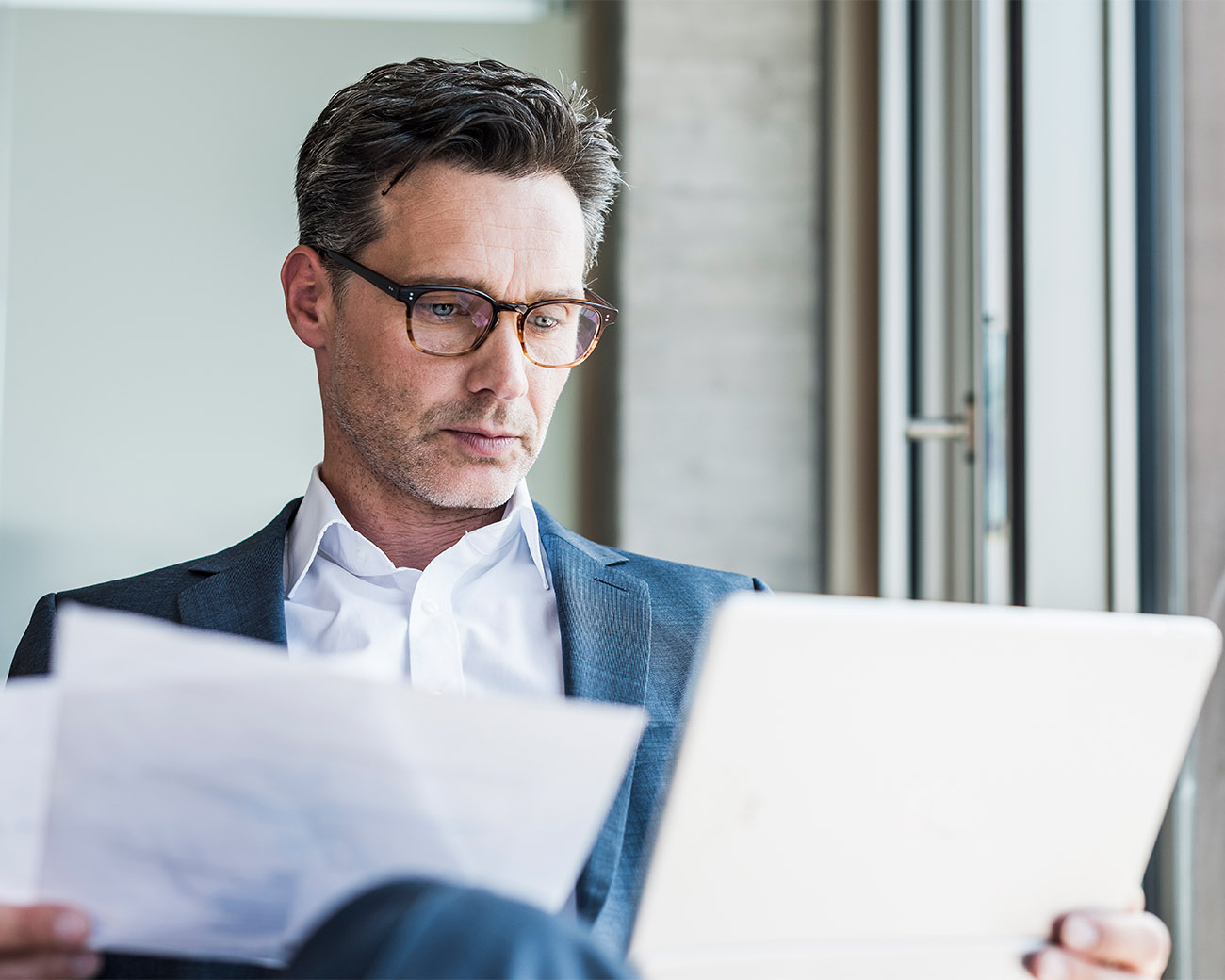 Man met bril heeft twee papieren in zijn hand en kijkt naar een tablet.