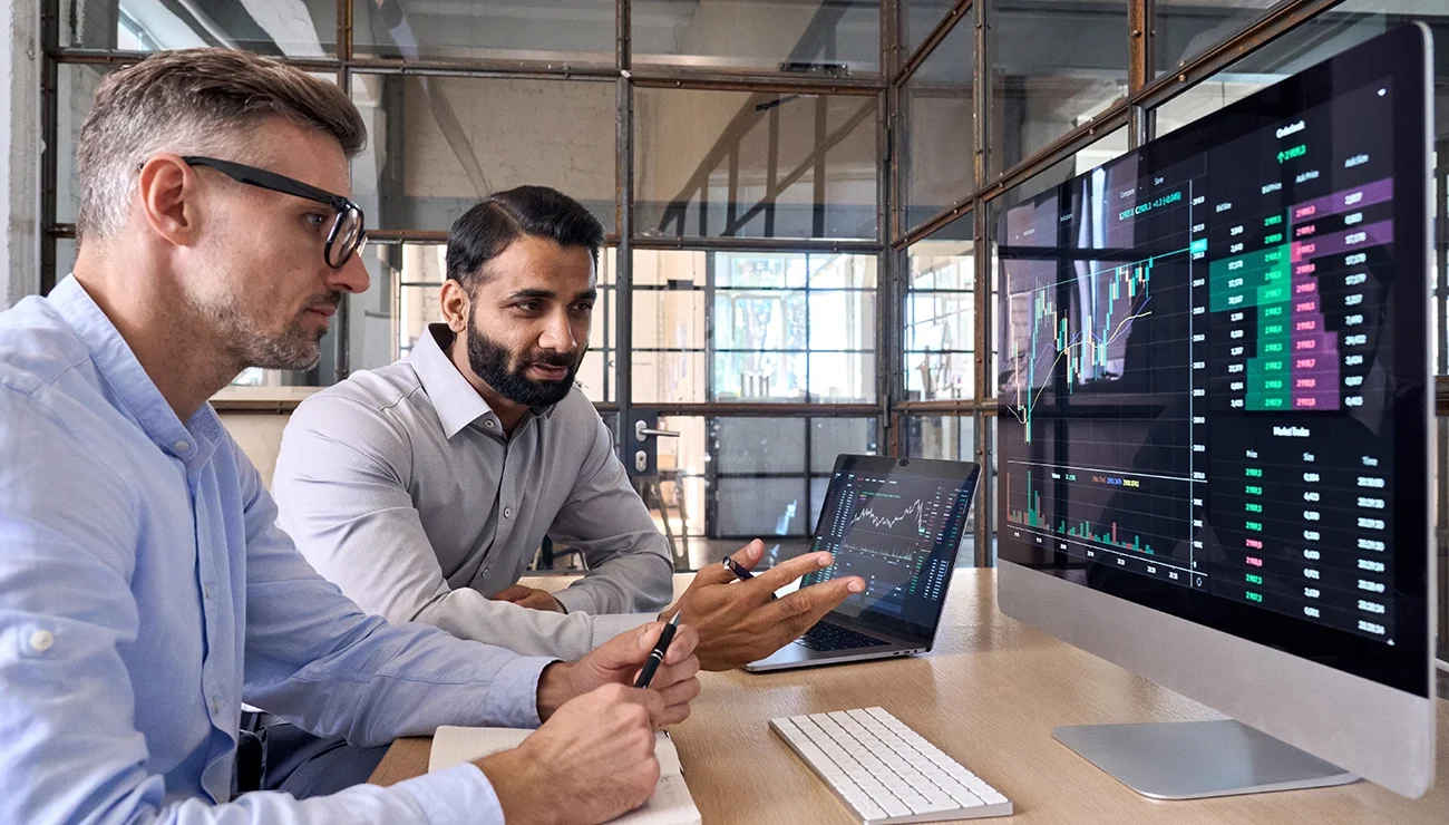 Twee personen bespreken handelsgrafieken en onderzoeksrapporten op een computerscherm