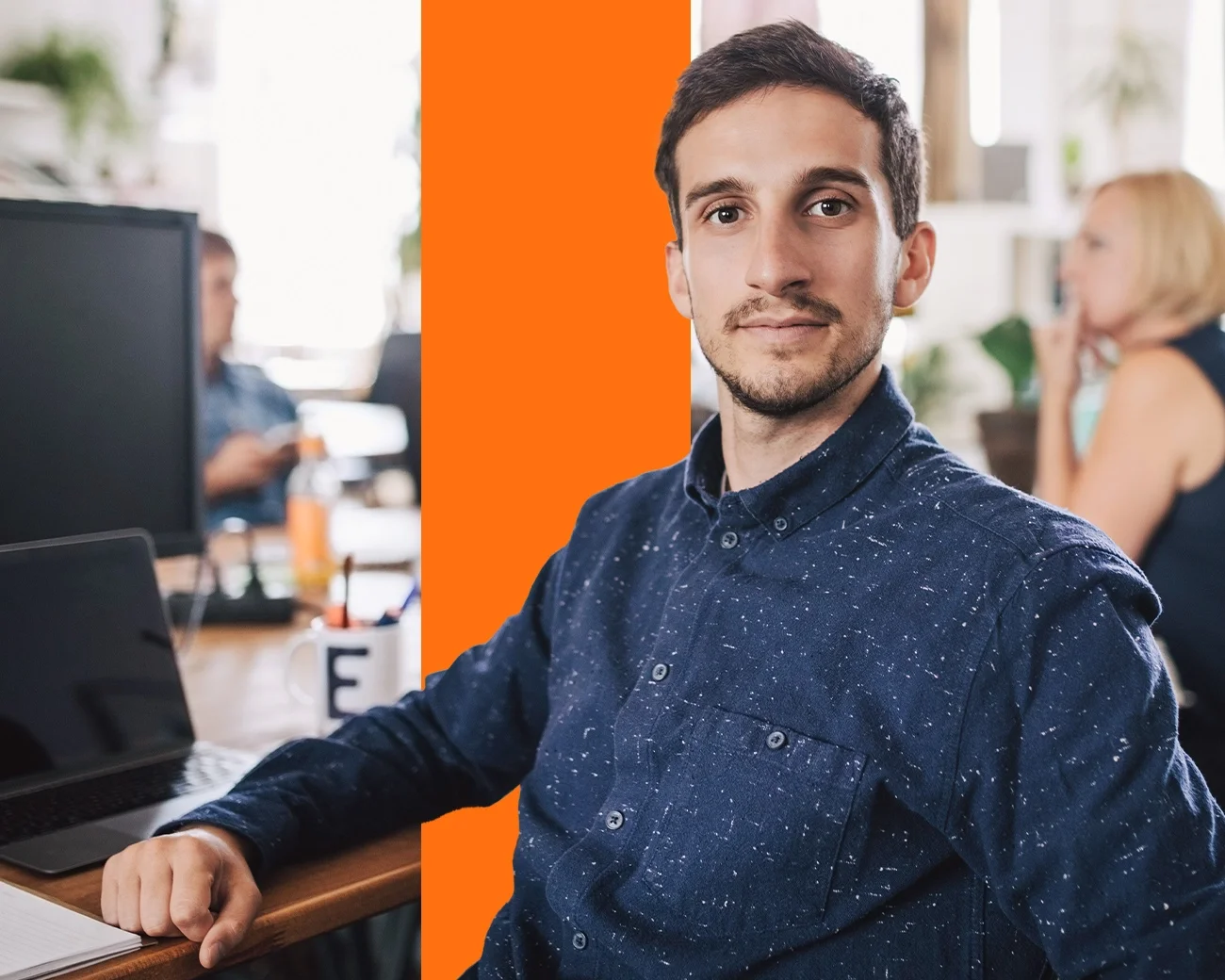 Jonge man met baard en blauw hemd zit achter bureau met oranje balk op de achtergrond