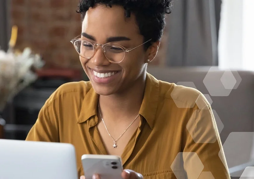 Vrouw met bril en okergele blouse met mobiel in haar hand kijkt naar laptopscherm