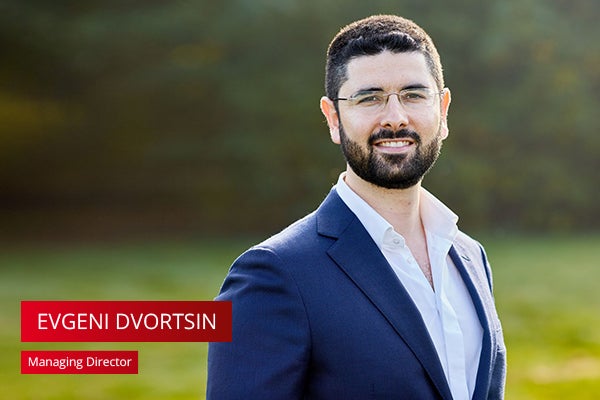 Man met kort bruin haar en baard met bril en donkerblauw pak staat buiten in groene omgeving. Tekst in rode balk: "Evgeni Dvortsin - Managing Director"
