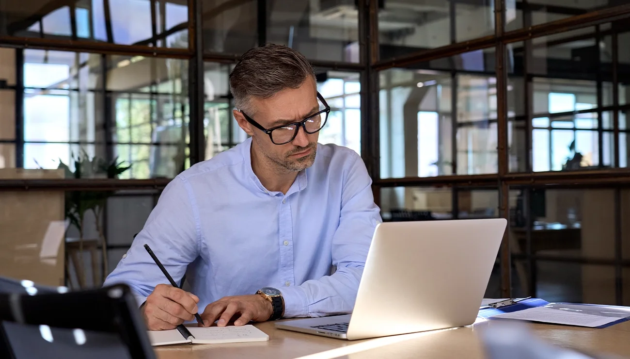 Man schrijft met pen in een notitieboek terwijl hij informatie van zijn laptop bekijkt