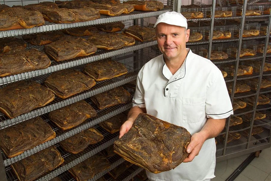 Hersteller der Rinner im Reiferaum hält ein Stück Südtiroler Speck g.g.A. vor Regalen mit weiteren Stücken