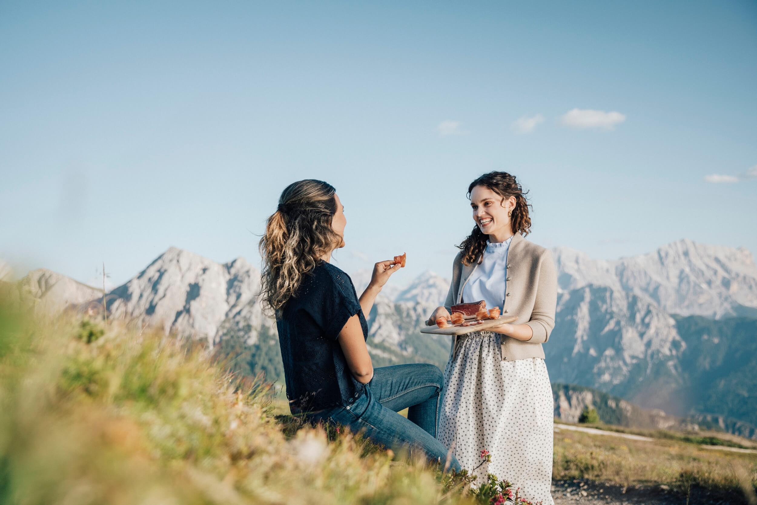 Zwei Frauen auf Almwiese essen Südtiroler Speck g.g.A. mit Bergen im Hintergrund