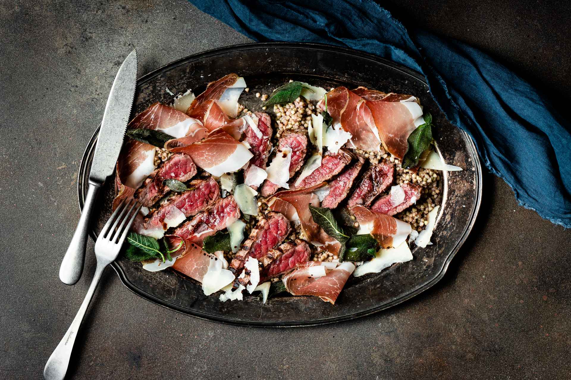 Tagliata wołowa ze speckiem z Południowego Tyrolu, kaszą gryczaną, płatkami parmezanu i liśćmi szałwii na ciemnym talerzu.