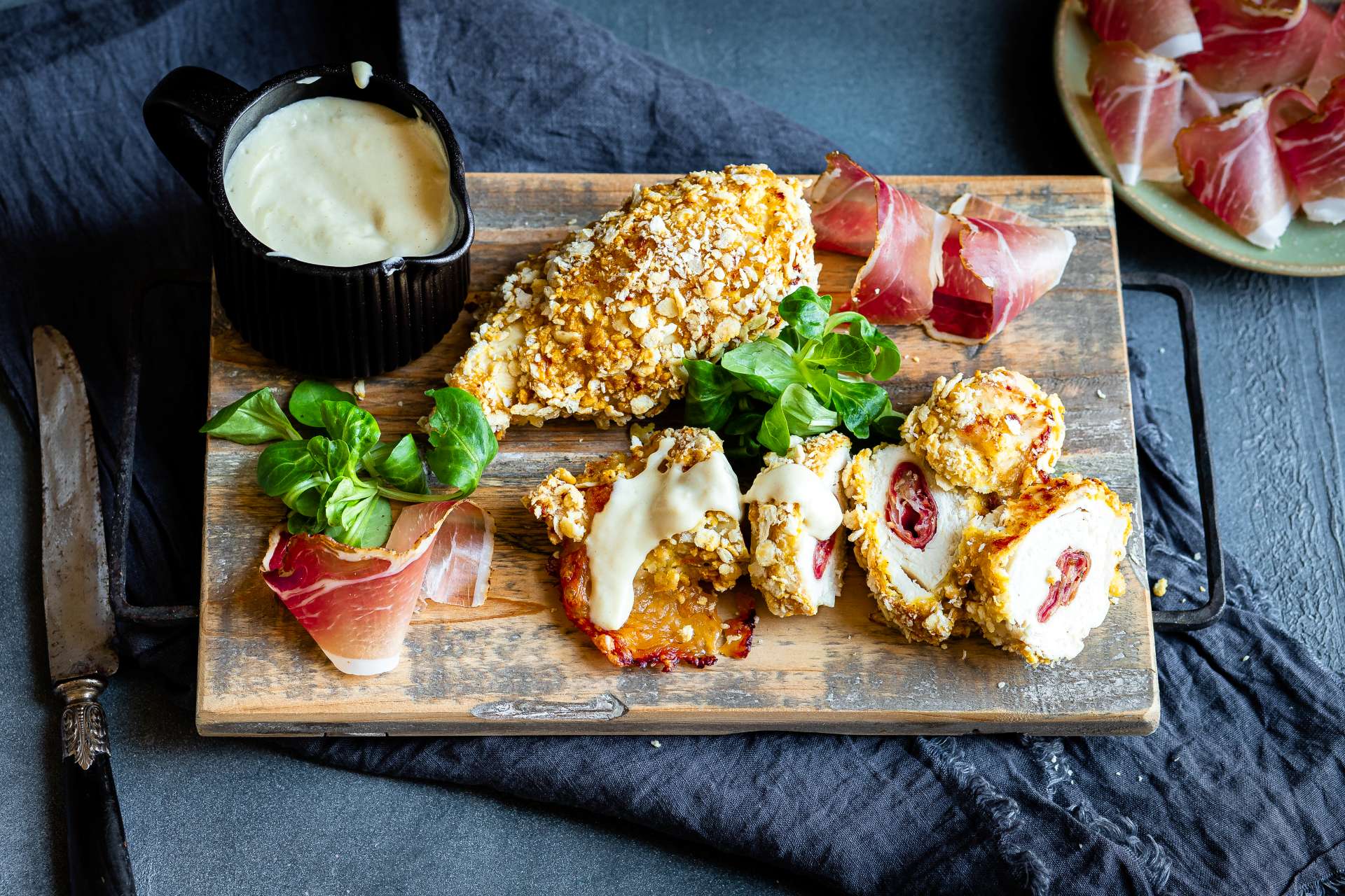 Panierter Hähnchen-Cordon-Bleu in Scheiben geschnitten, gefüllt mit Südtiroler Speck g.g.A. und Mozzarella, serviert auf einem Brett mit Senfsauce sowie Südtiroler Speck g.g.A. daneben.