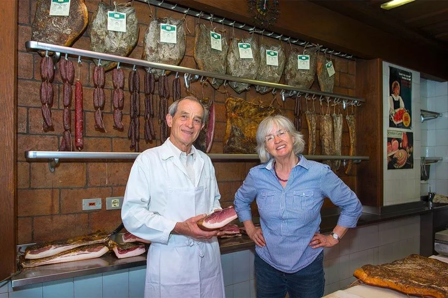 Produzenten der Metzgerei Gstör im Innenraum vor Wand mit Wurstwaren und Südtiroler Speck g.g.A., eine hält ein Stück