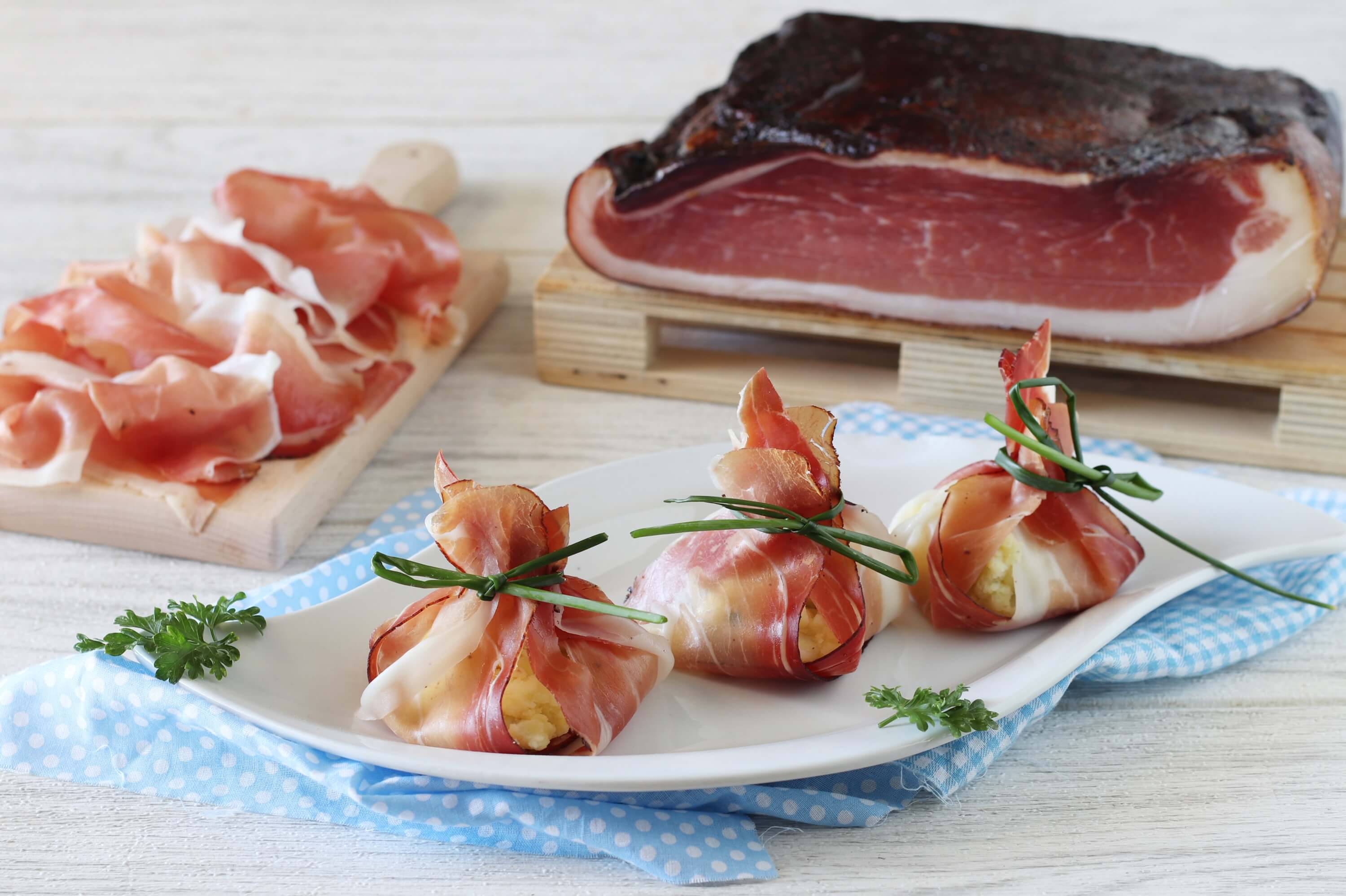 Potato-filled Speck Alto Adige PGI purses, wrapped in slices of Speck Alto Adige PGI and tied with chives, served on a white plate with whole and sliced Speck Alto Adige PGI in the background.