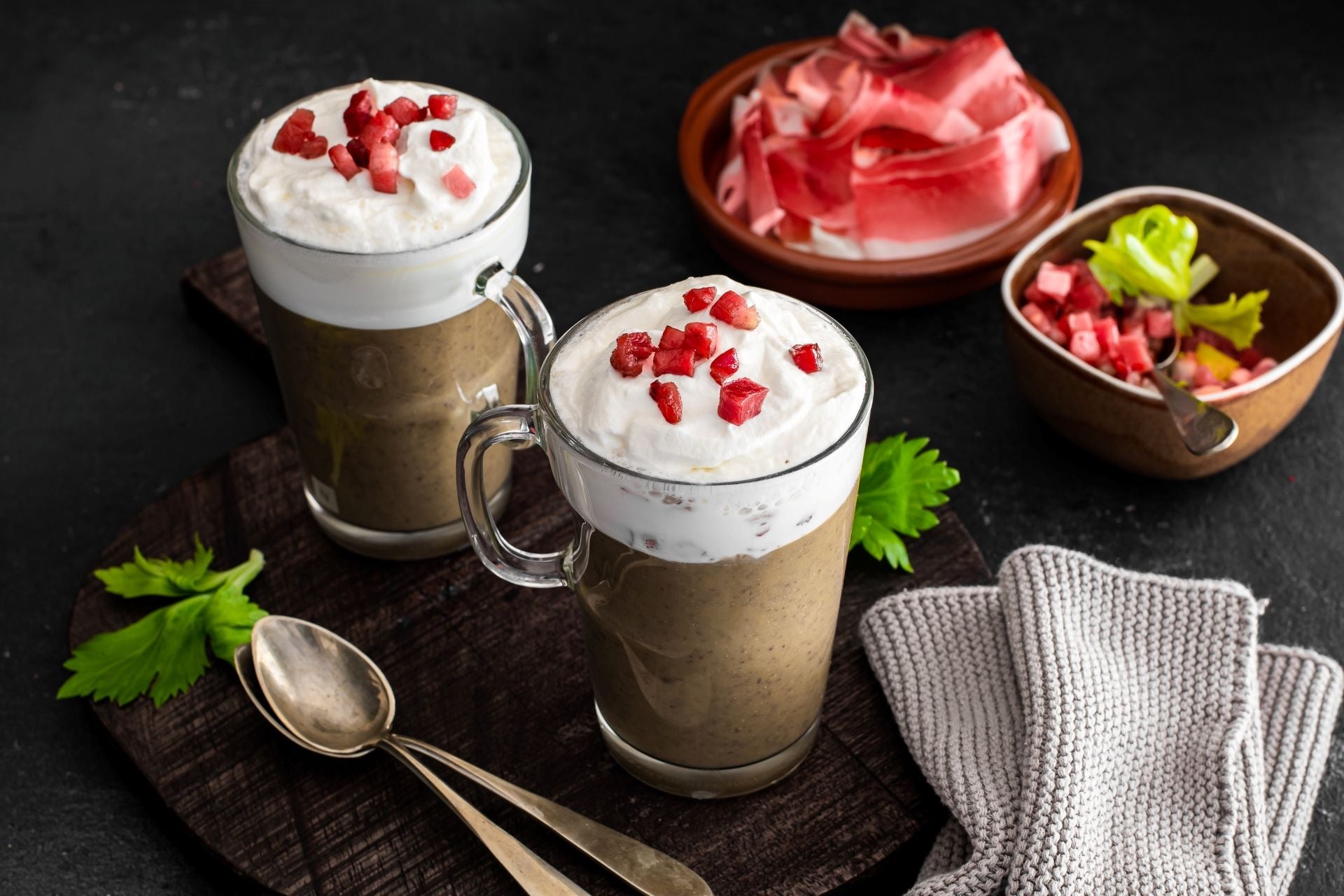 Lentil cappuccino served in glass cups, topped with creamy foam and crispy Speck Alto Adige PGI, presented on a dark surface.