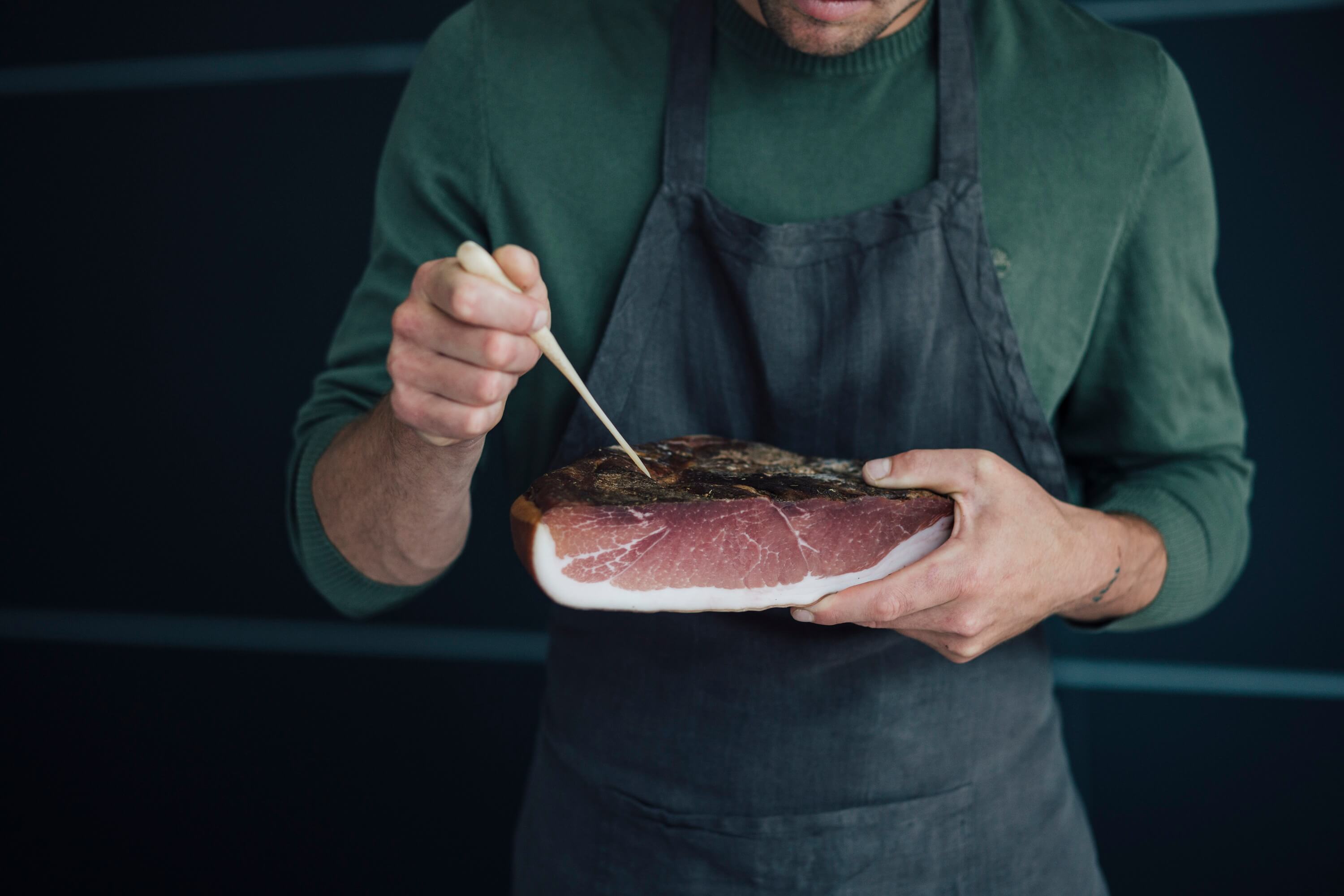 Hersteller prüft Speck Südtiroler Speck g.g.A. mit Nadel, hält Stück Speck in Händen.