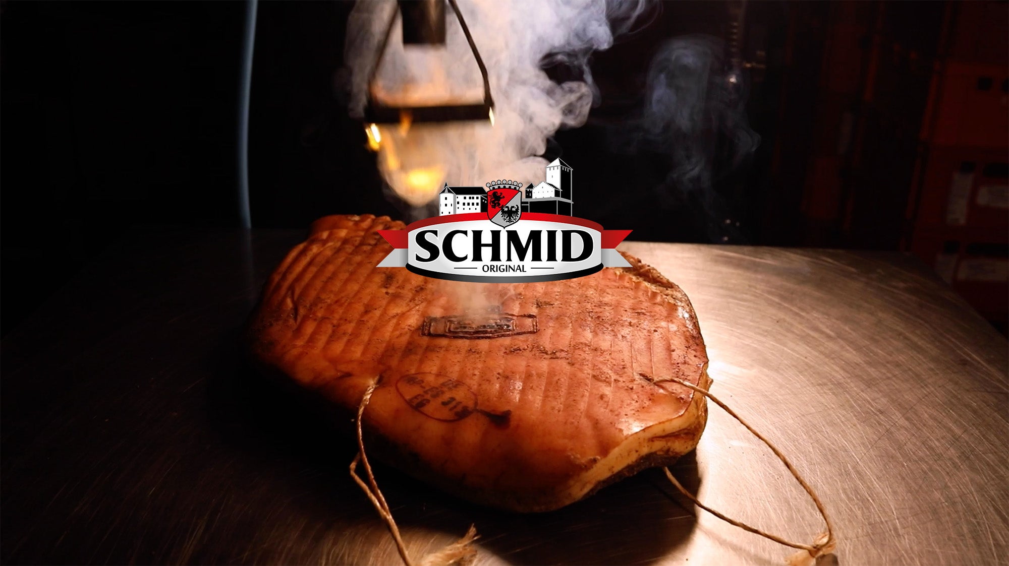 Piece of Speck Alto Adige PGI being smoked with visible smoke and Schmid Original logo in foreground