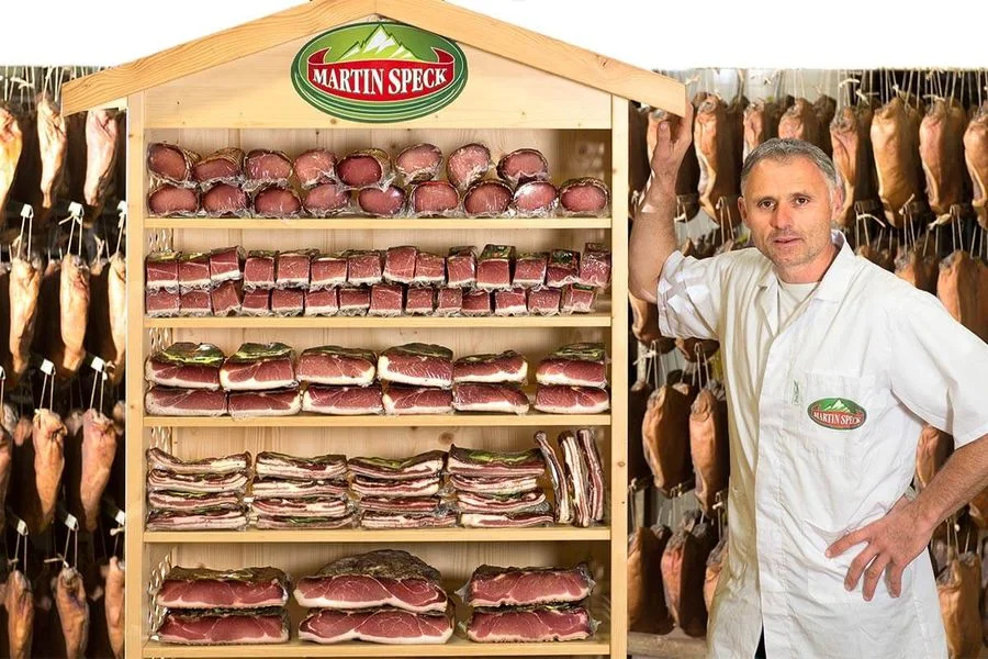 Martin Dipauli im Innenraum neben Regal mit Südtiroler Speck g.g.A., im Hintergrund hängende Stücke