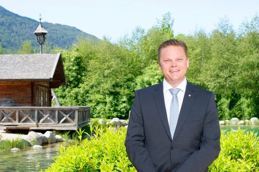 Karl Christian Handl posiert im Freien vor Holzgebäude und Berglandschaft im Hintergrund