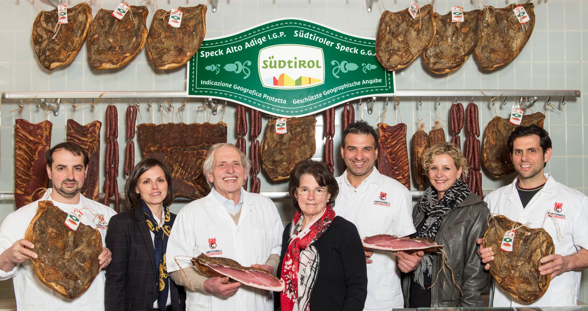 Il team della macelleria Christanell in posa con diversi pezzi di Speck Alto Adige IGP, insegna del marchio di qualità Alto Adige sulla parete
