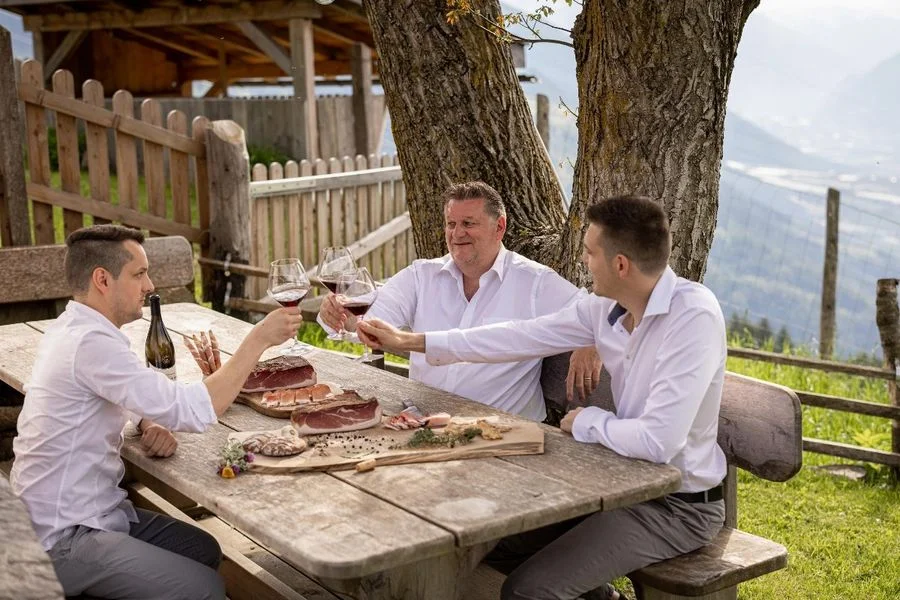 Familie Moser stößt am Tisch im Freien mit Rotwein an, Südtiroler Speck g.g.A. und Brot auf dem Brett