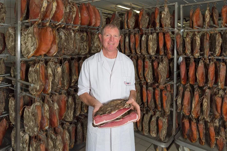 Produttore tiene in mano Speck Alto Adige IGP, sullo sfondo speck appeso in stagionatura.
