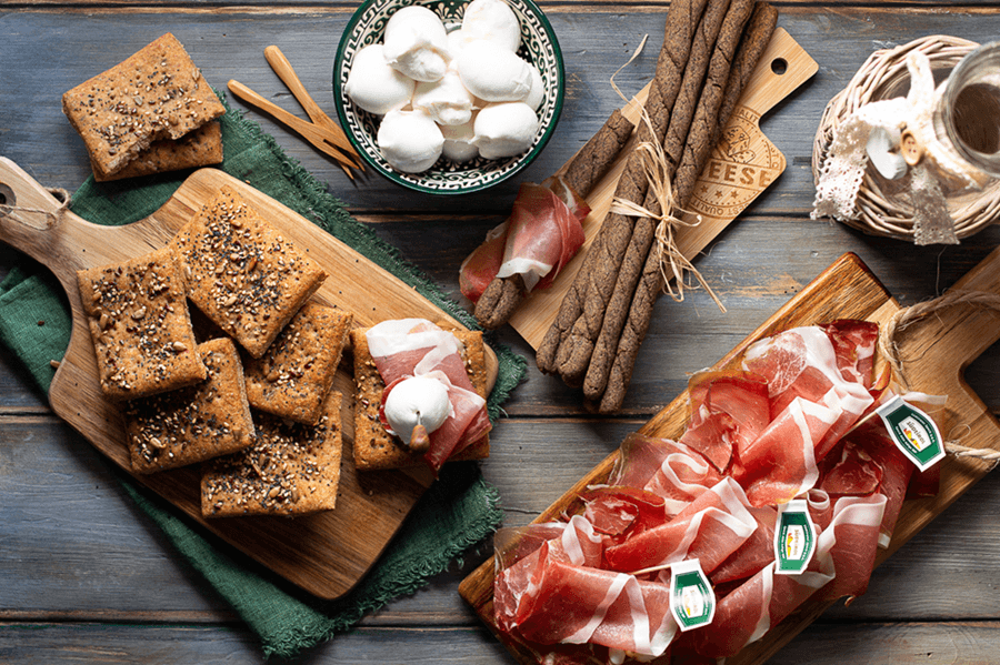 "Schiacciatine" complètes aux graines et gressins de sarrasin au Speck Alto Adige IGP, servis sur planches en bois avec des bouchées de mozzarella.