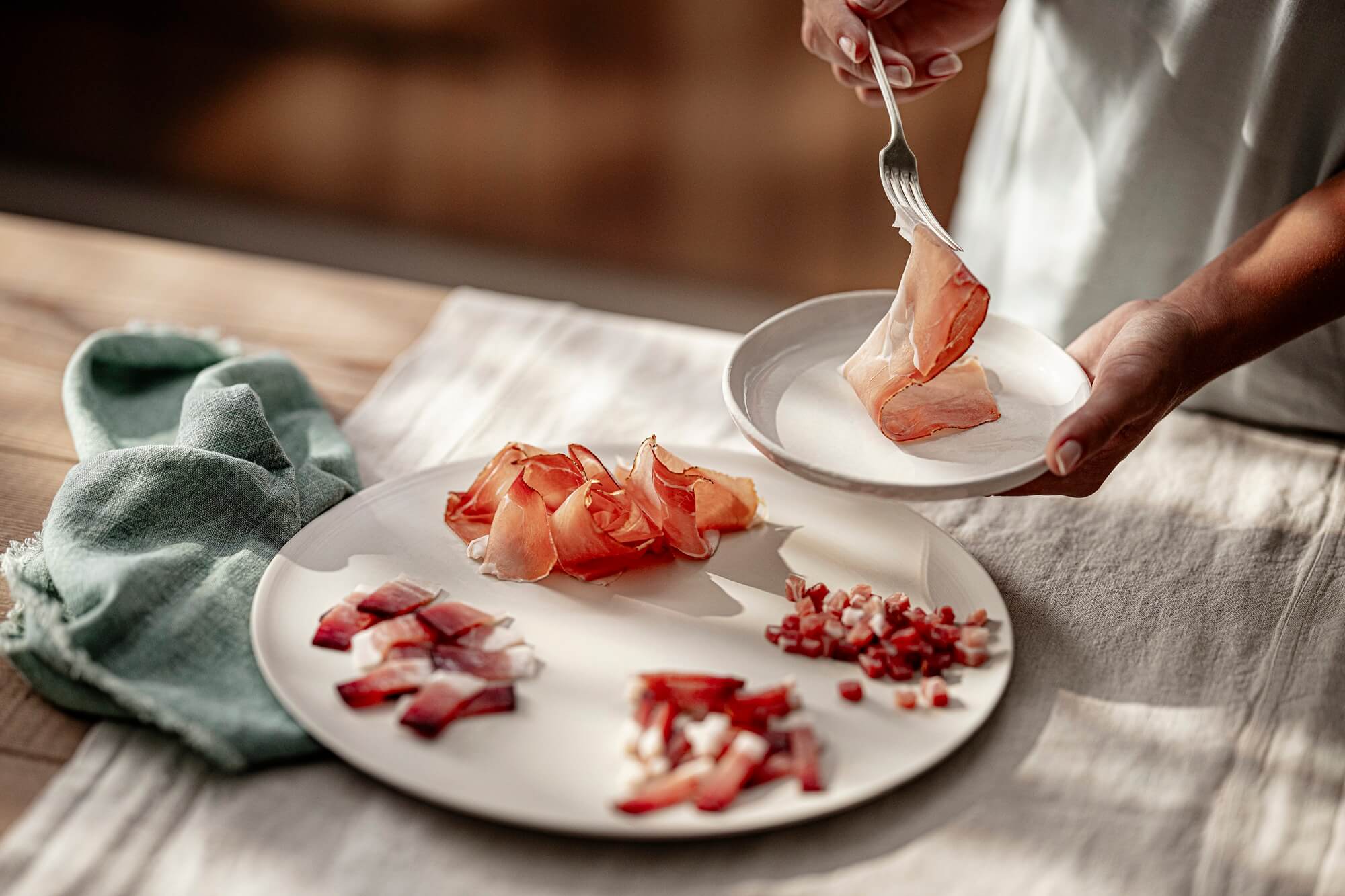 Verschiedene Schnitte von Südtiroler Speck g.g.A. auf Teller, Scheibe wird mit Gabel genommen.