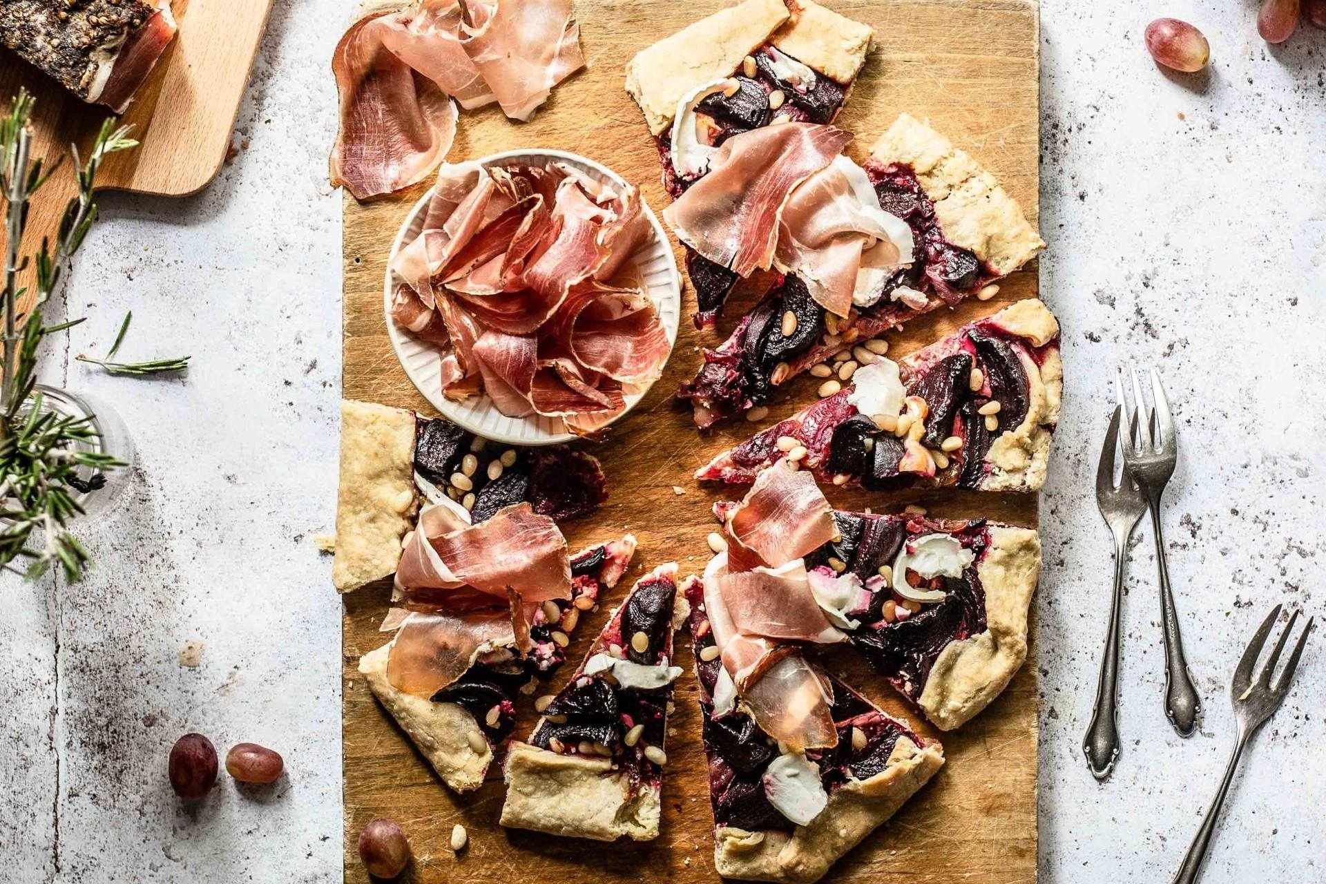 Galette à la betterave avec oignons glacés, fromage de chèvre et Speck du Tyrol du Sud IGP, servie sur une planche rustique.