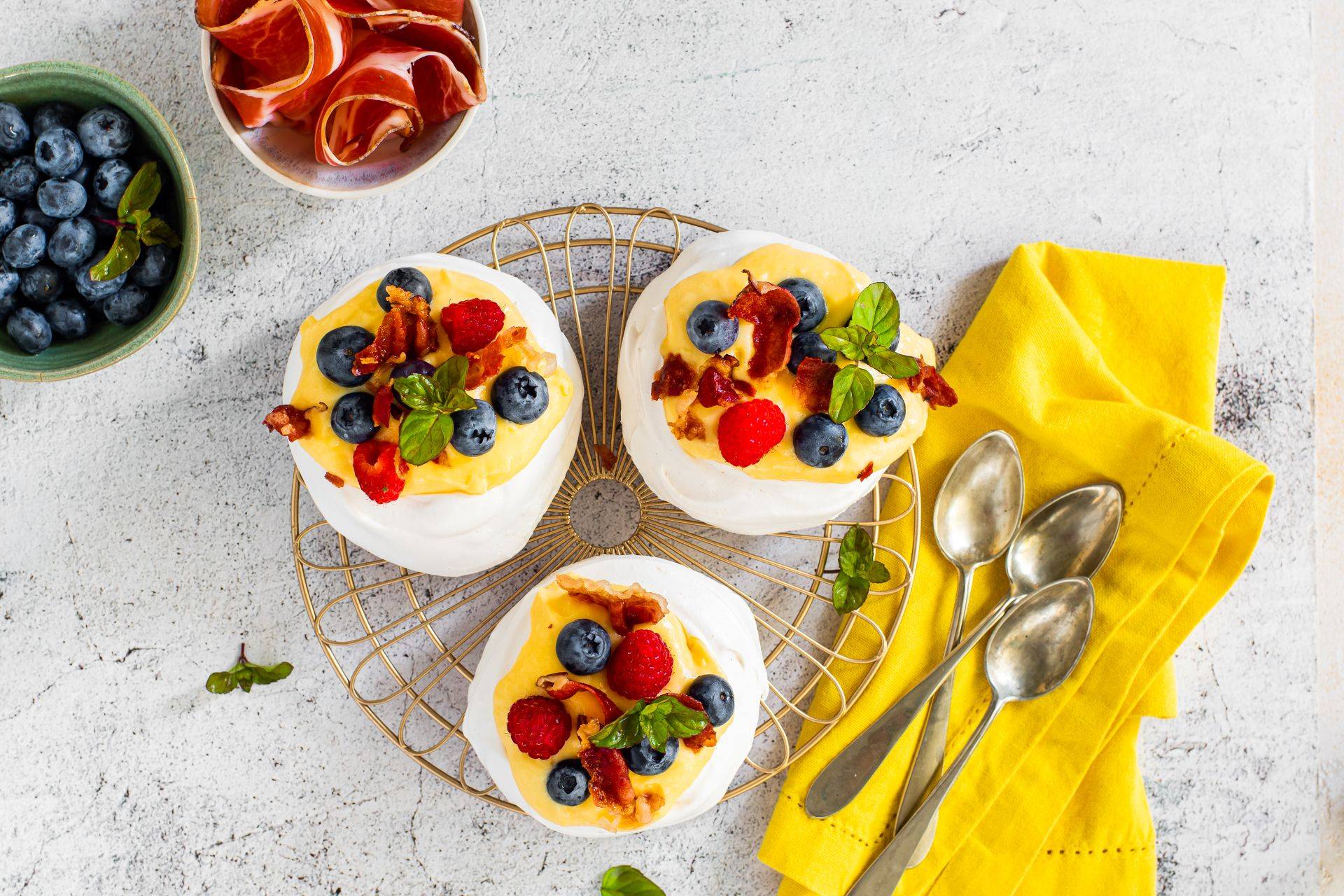 Mini-Pavlovas aus Baiser, gefüllt mit cremiger Vanillecreme, garniert mit knusprigem Speck Alto Adige IGP, Heidelbeeren, Himbeeren und frischen Minzblättern, auf einem goldenen Gitter serviert.