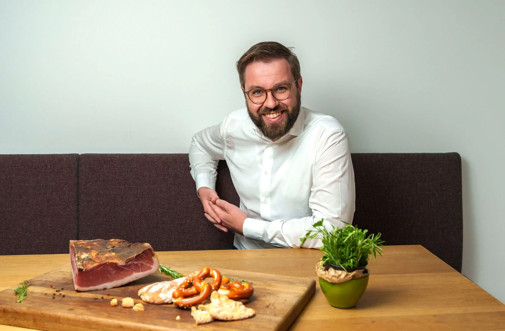 Lukas Pfitscher posing at a table with Speck Alto Adige PGI and pretzels on a wooden board with herbs in a pot