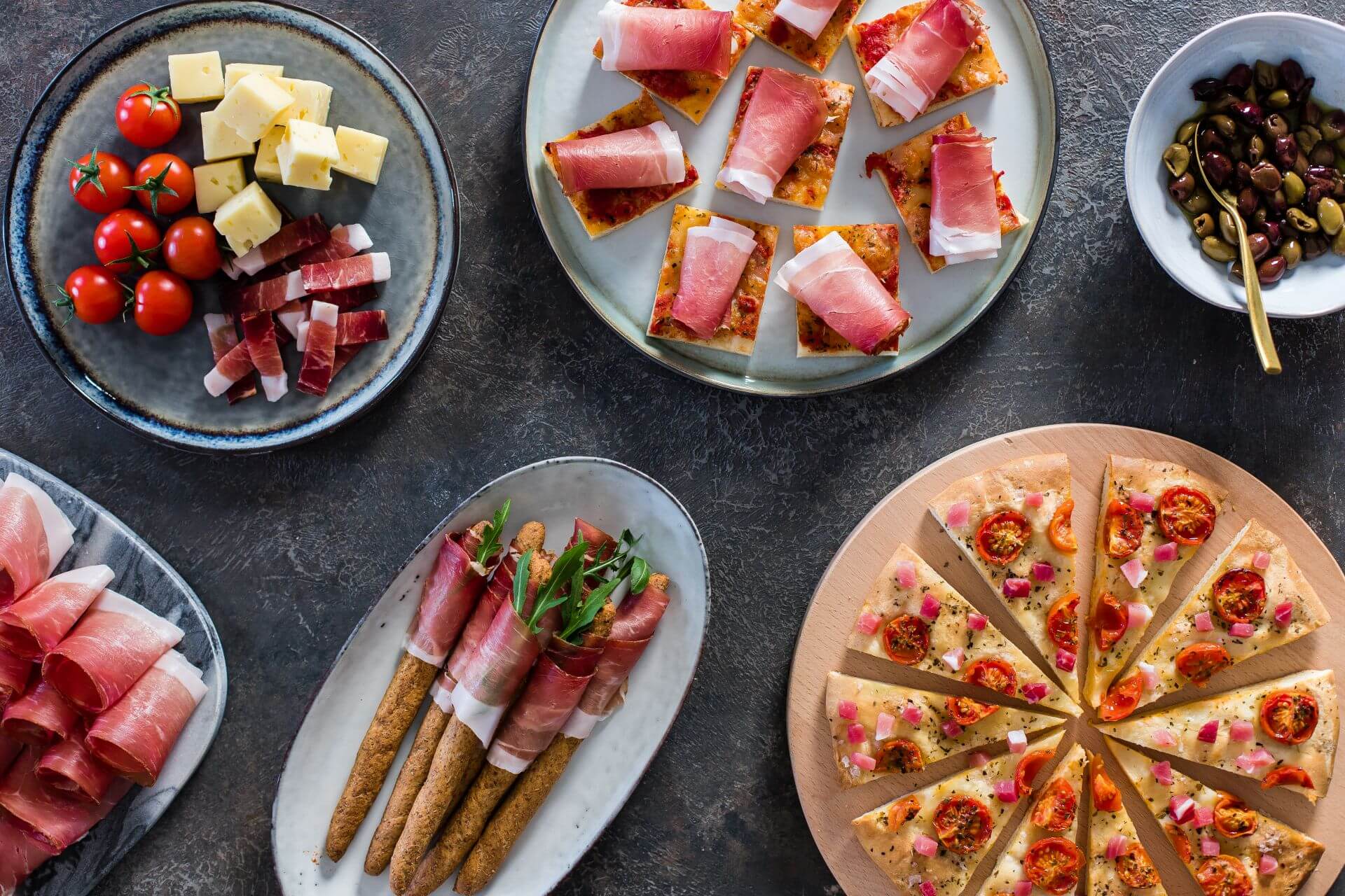 Verschiedene Häppchen mit Südtiroler Speck g.g.A. auf Tellern mit Käse, Tomaten und Oliven.