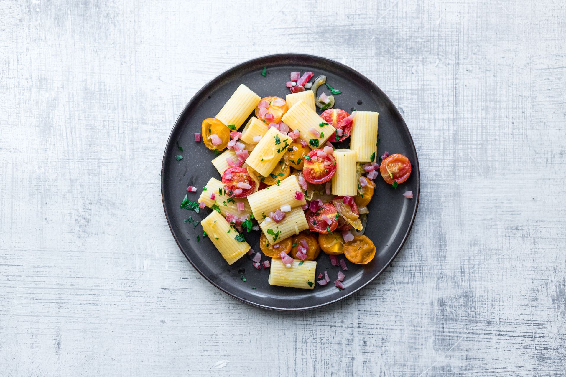 Maccheroni with South Tyrolean Speck PGI, cherry tomatoes and fresh herbs, served on a dark plate, top view.