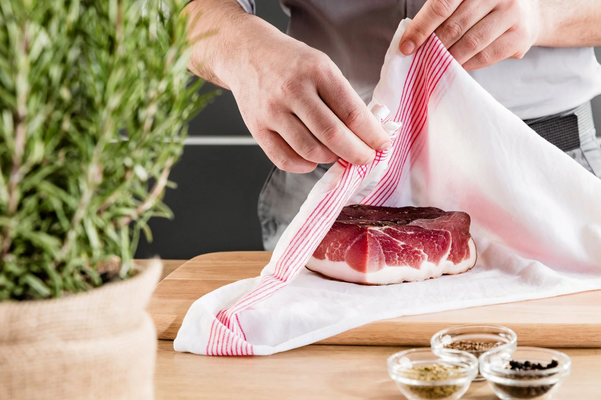 Person wickelt Stück Südtiroler Speck g.g.A. mit Küchentuch auf Brett, daneben Gewürze und Rosmarin.