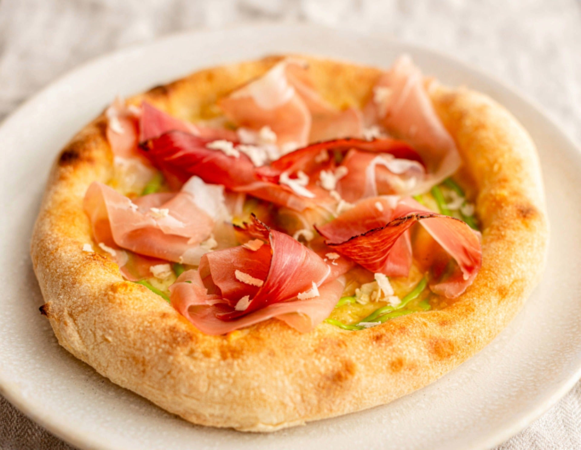 Hausgemachte Pizza auf weißem Teller mit hohem, goldbraunem Rand, belegt mit Südtiroler Speck Alto Adige g.g.A., Mayonnaise und Schnittlauch.