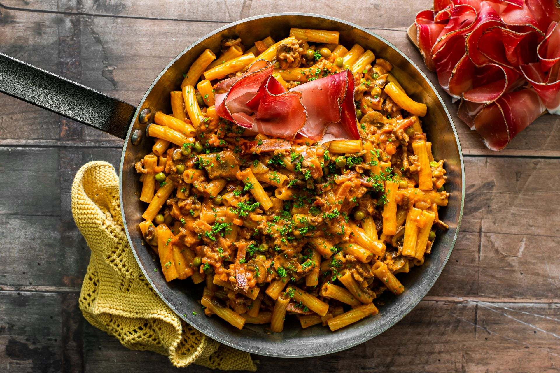 Kurze Pasta in einer herzhaften Sauce mit Fleisch und Gemüse, verfeinert mit Speck Alto Adige IGP und frischen Kräutern, in einer Pfanne angerichtet.