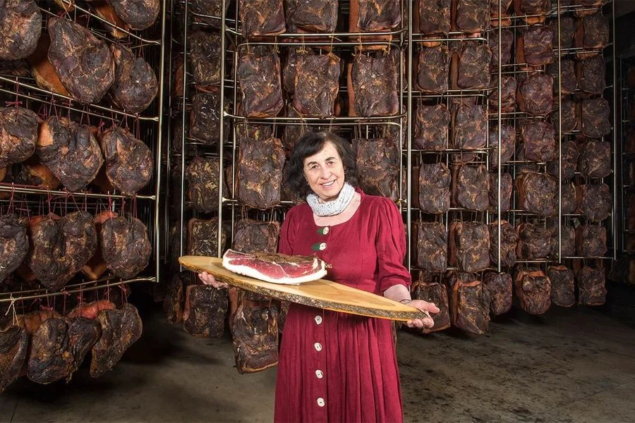 Inge Rauch im Reiferaum hält Brett mit Südtiroler Speck g.g.A. vor Regalen voller hängender Stücke