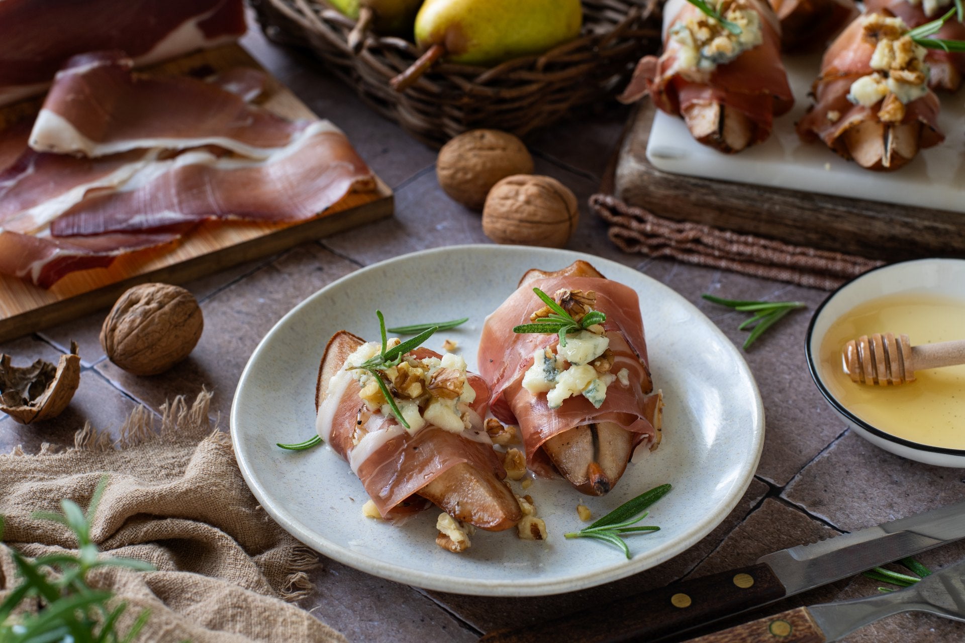Gebackene Birnen, umwickelt mit Südtiroler Speck, gefüllt mit Gorgonzola und Walnüssen, auf einem Teller serviert, mit Honig beträufelt und mit Rosmarin garniert.