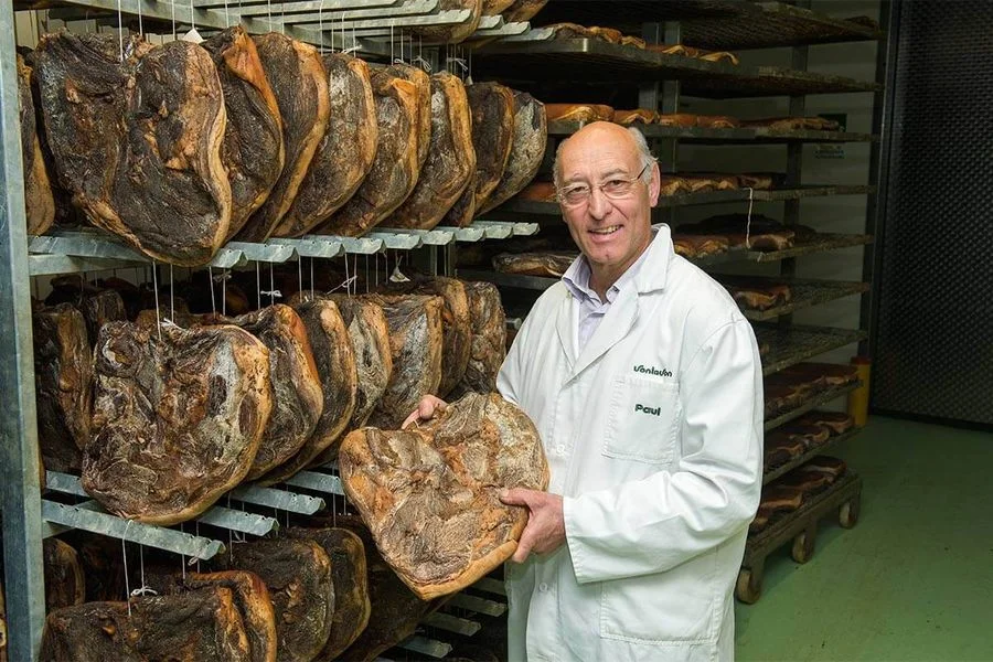 produttore Paul Vontavon in locale di stagionatura tiene in mano un pezzo di Speck Alto Adige IGP, accanto altri pezzi appesi