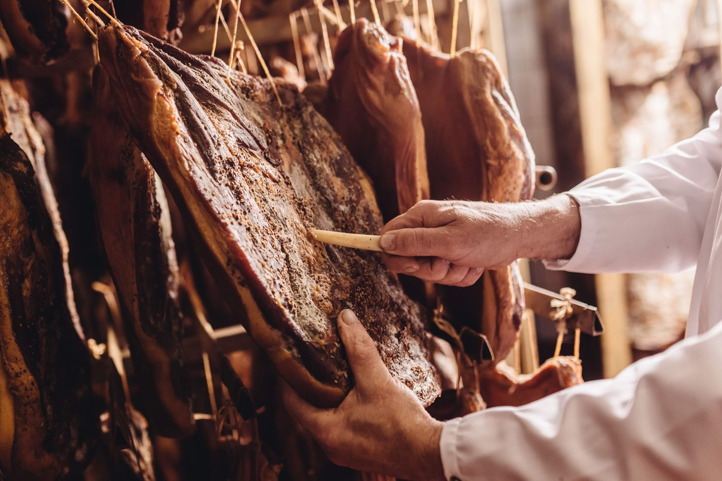 Hände mit Holzwerkzeug prüfen Oberfläche von Südtiroler Speck g.g.A. im Reiferaum