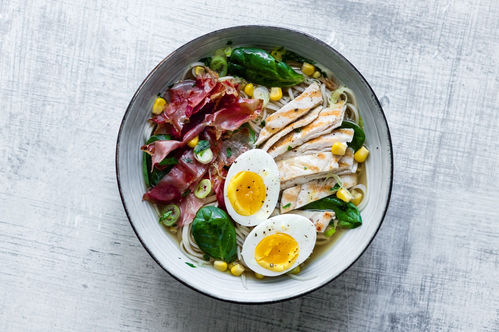 Ramen au speck du Tyrol du Sud IGP, œuf, poulet, maïs et épinards, servi dans un bol, vue de dessus.