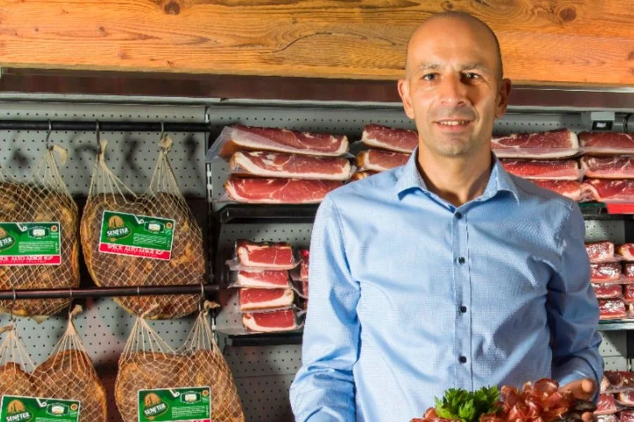 Hersteller der Senfter steht im Innenraum vor Regalen mit Südtiroler Speck g.g.A. und hält ein Brett mit Speck