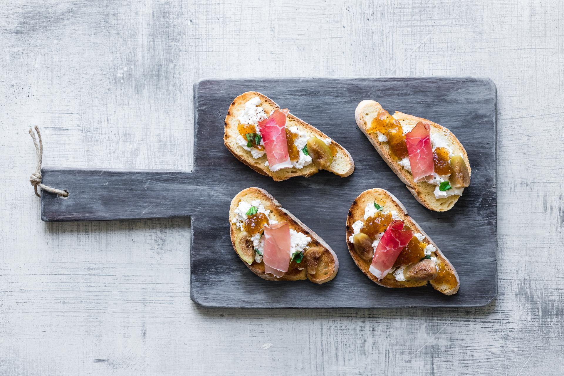 Bruschette con ricotta, Speck Alto Adige IGP e fichi, servite su un tagliere in legno scuro.