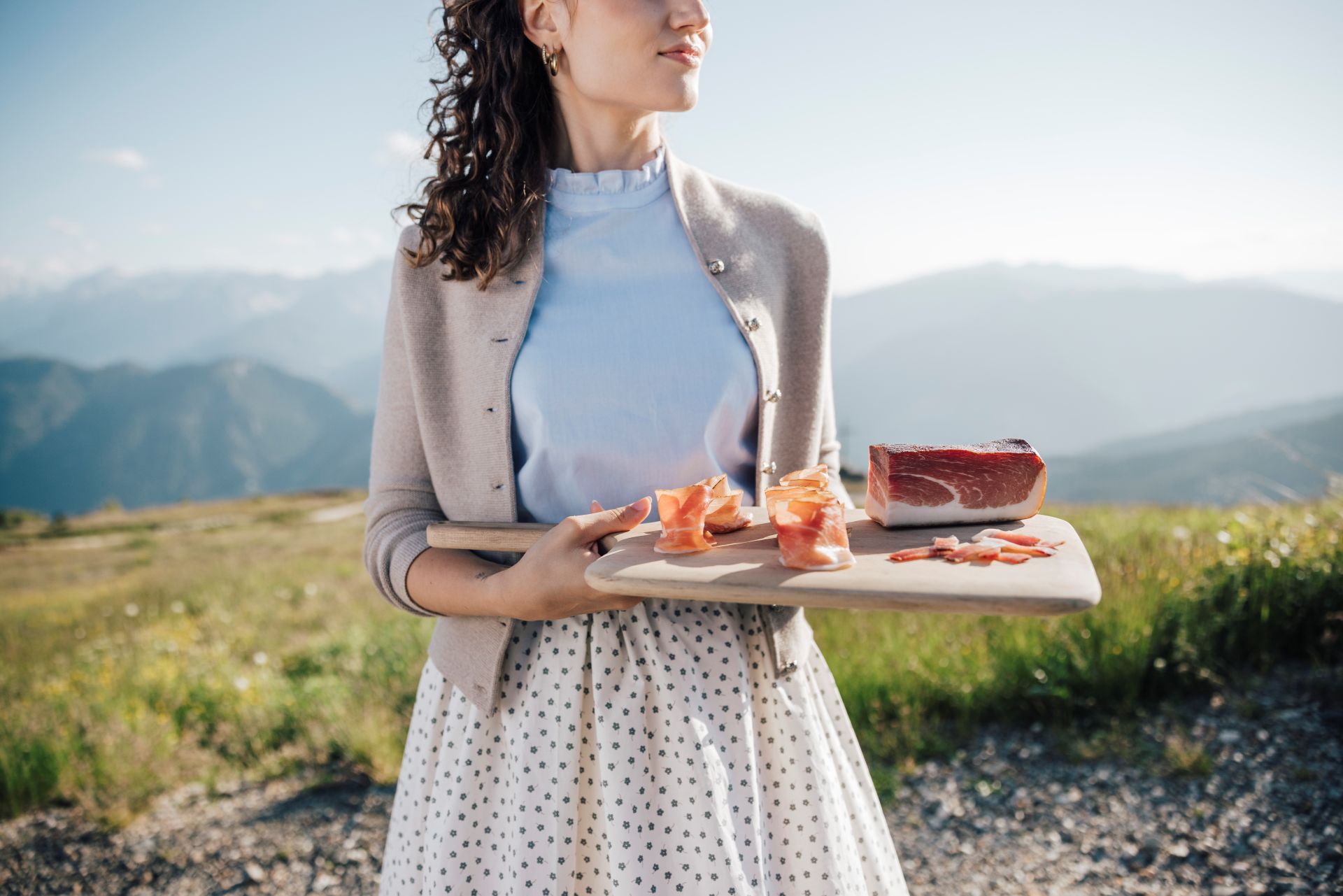 Frau hält Brett mit Speck Alto Adige g.g.A. und Scheiben vor Berglandschaft im Hintergrund
