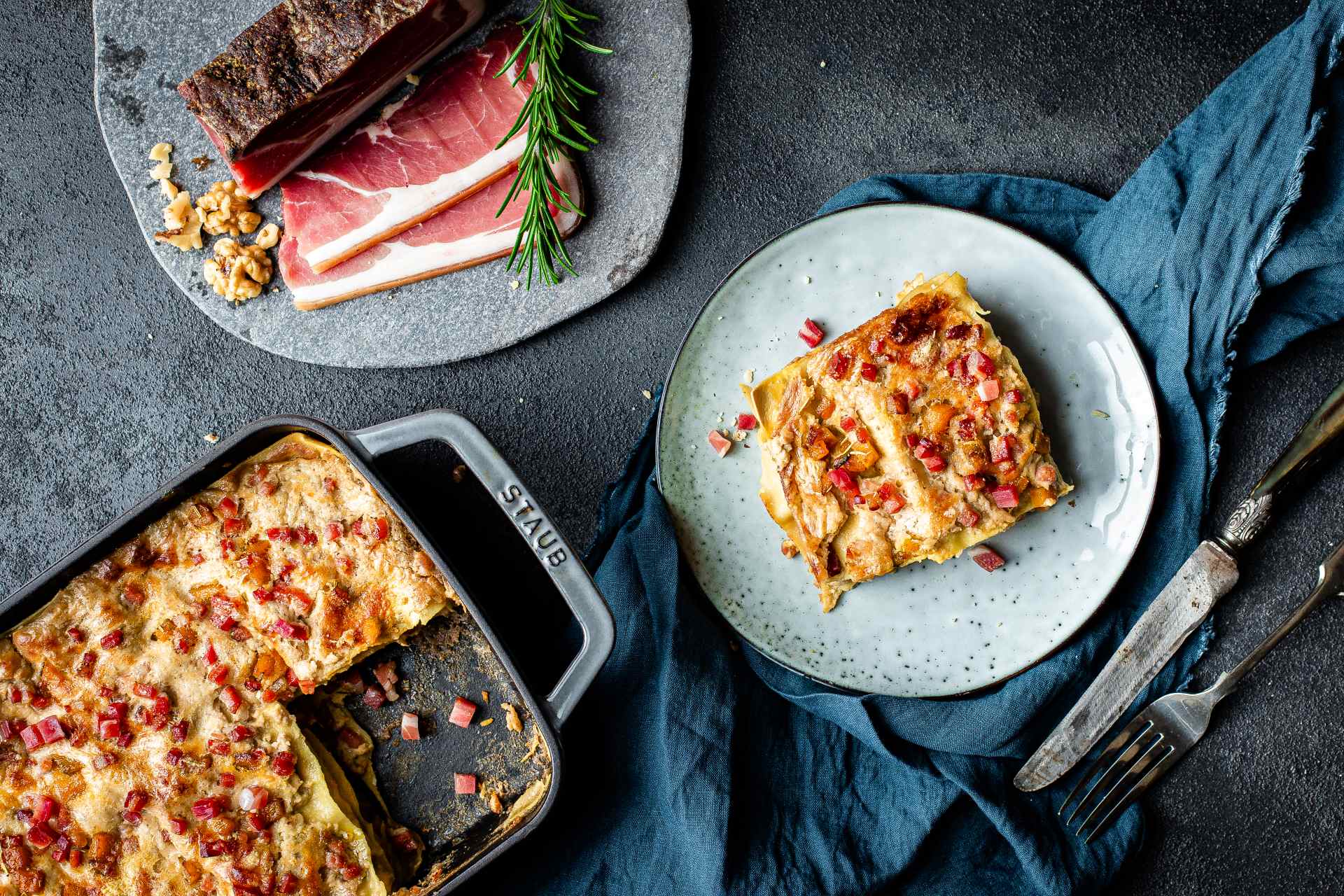 Gratinierte Lasagne mit Südtiroler Speck g.g.A.-Würfeln, Walnusscreme und Kürbis; daneben eine Form mit weiteren Lasagne und separaten Scheiben Südtiroler Speck g.g.A.