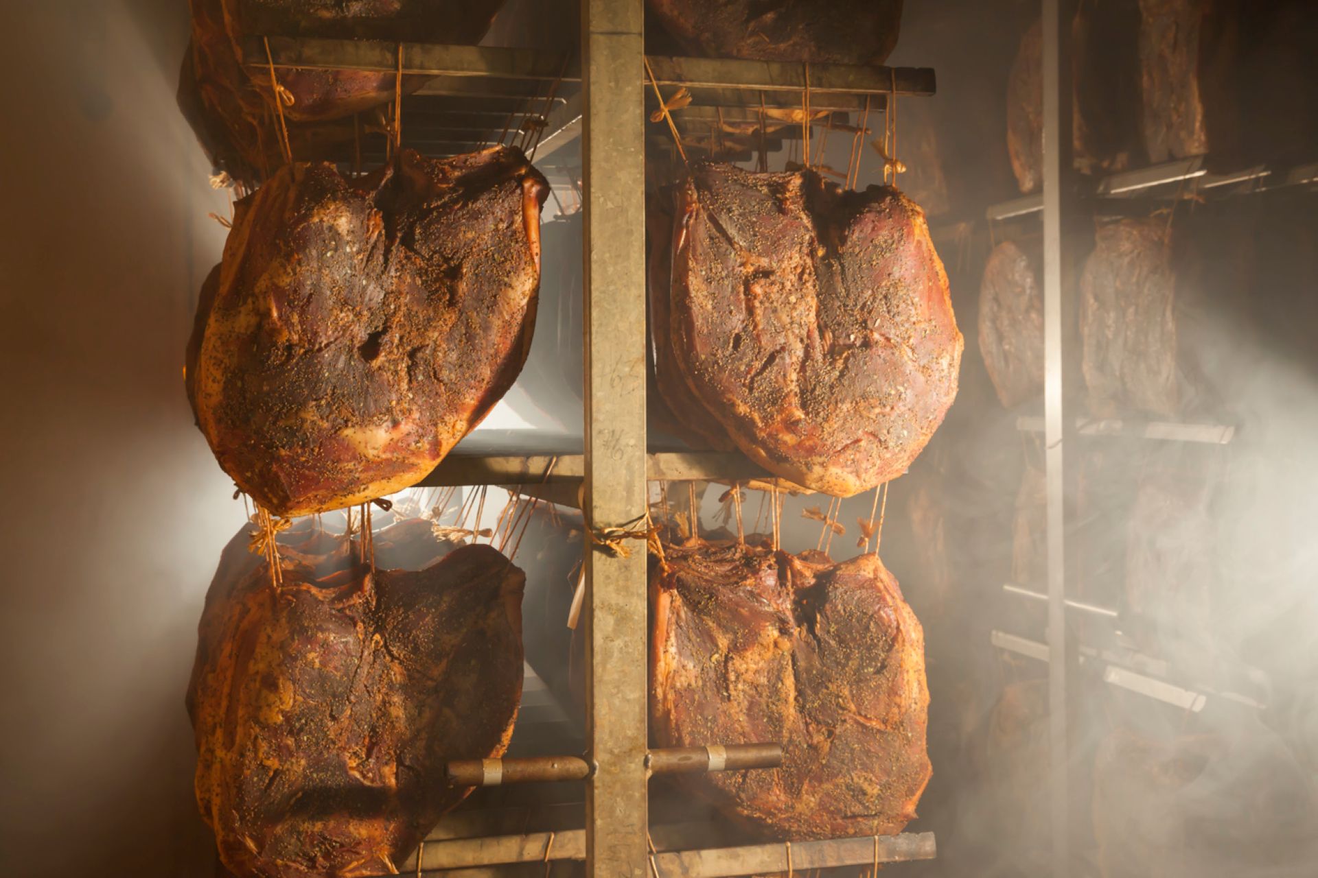 Mehrere Stücke Südtiroler Speck g.g.A. hängen zum Räuchern in einer Räucherkammer.