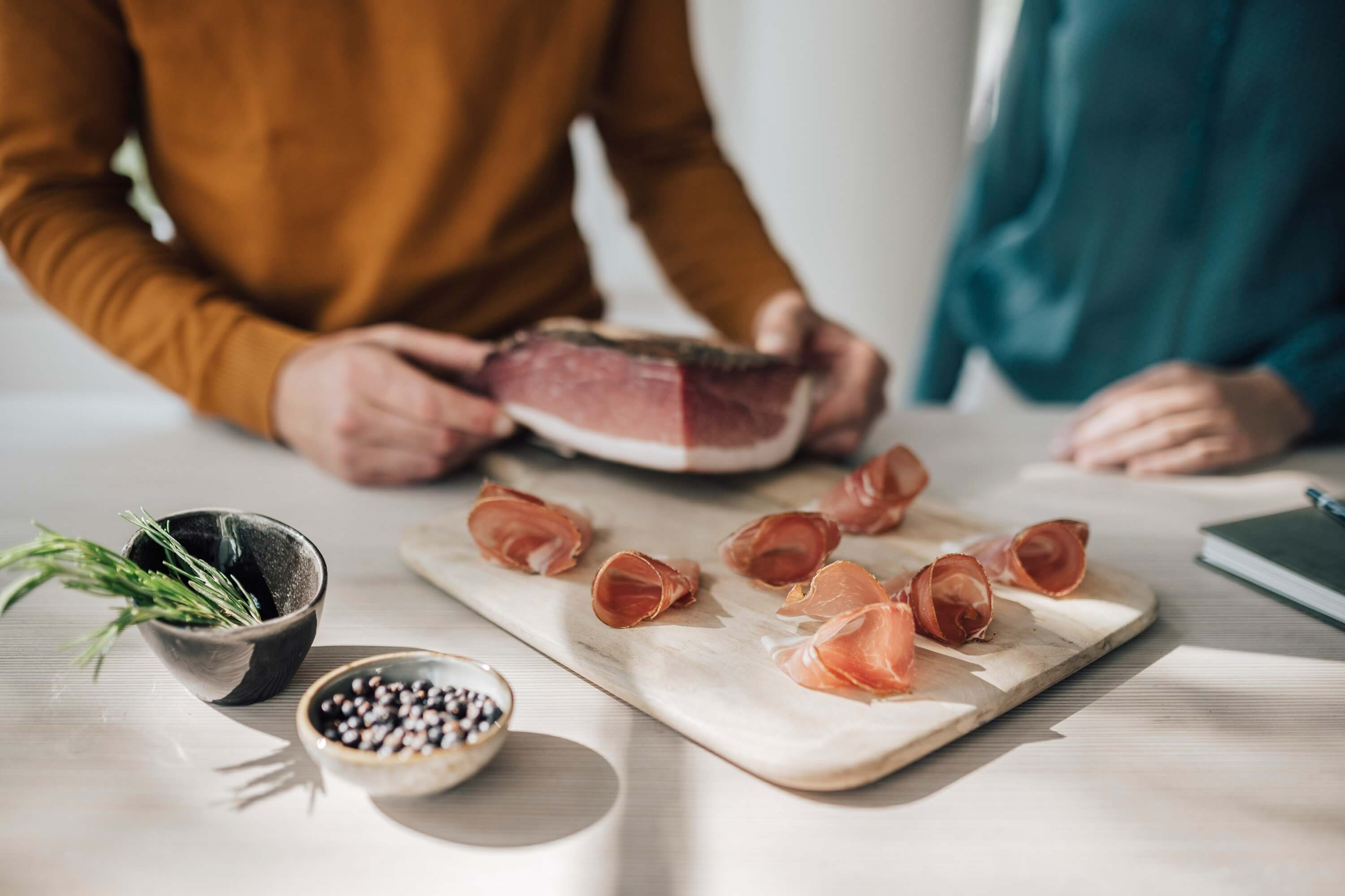 Zwei Personen mit Stück und Scheiben Südtiroler Speck g.g.A. auf Holzbrett, Gewürze und Rosmarin daneben.