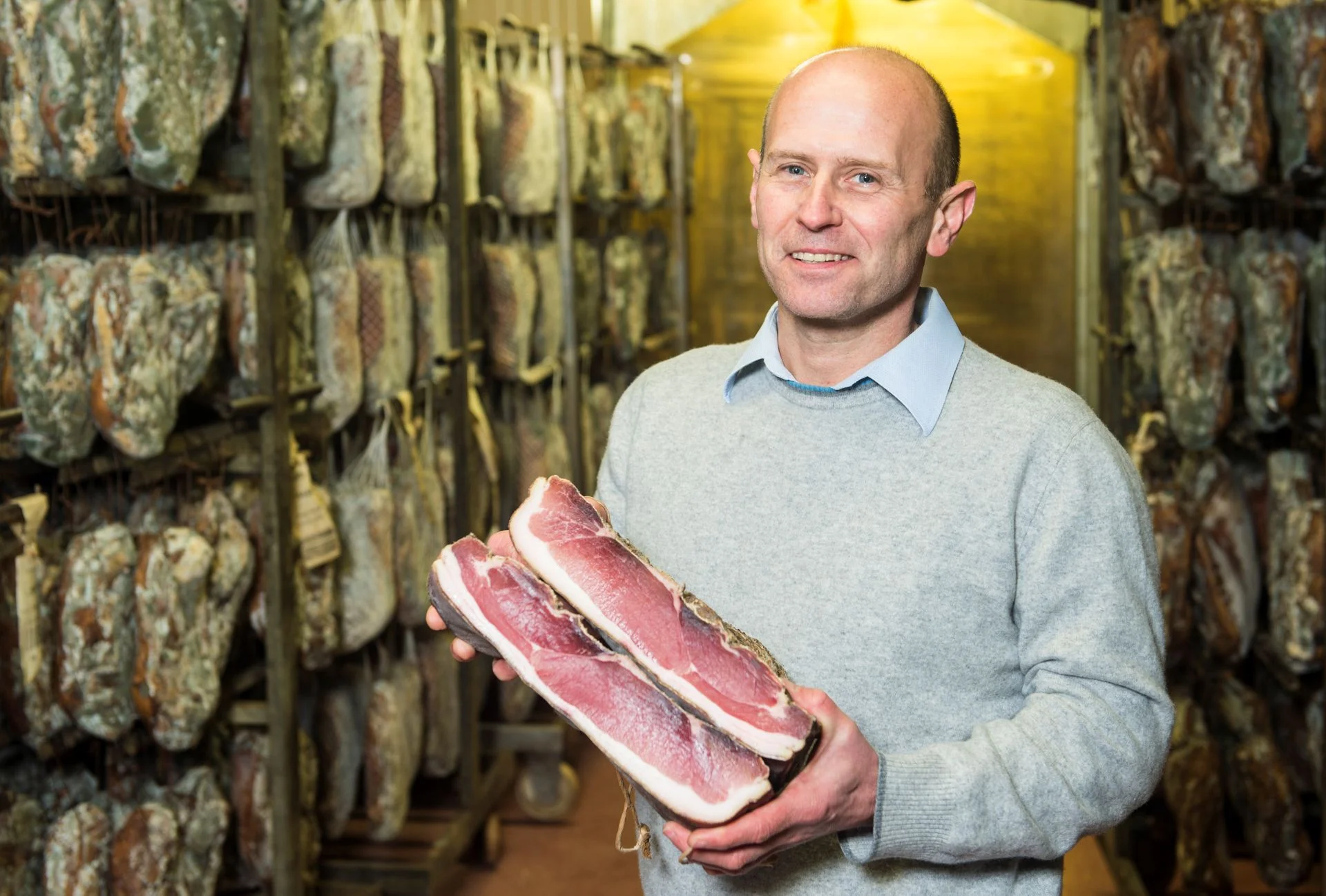 Peter Siebenförcher posing with two halved pieces of Speck Alto Adige PGI in a curing room