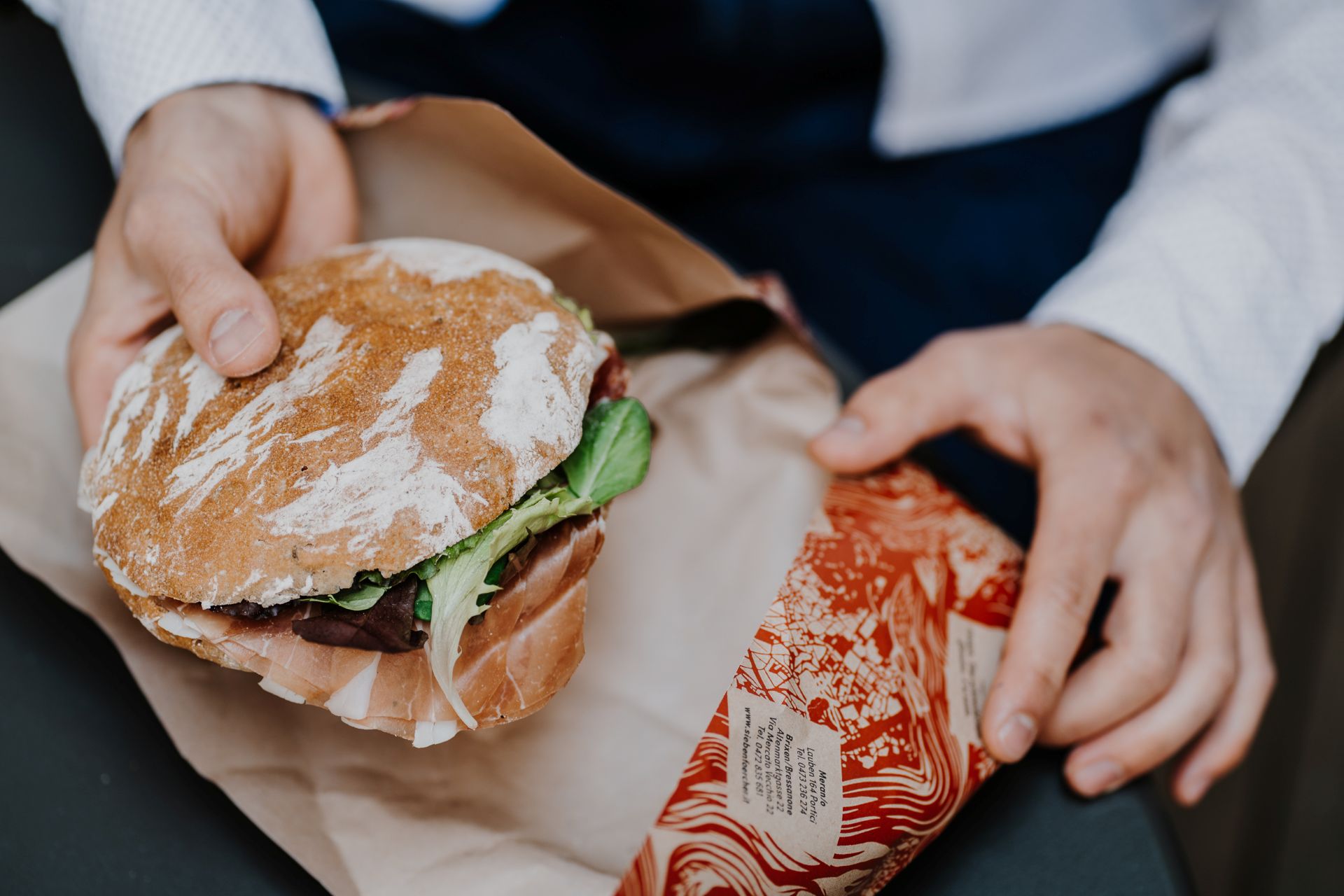 Hand hält belegtes Brot mit Südtiroler Speck g.g.A. und Salat, wird in Papier eingewickelt