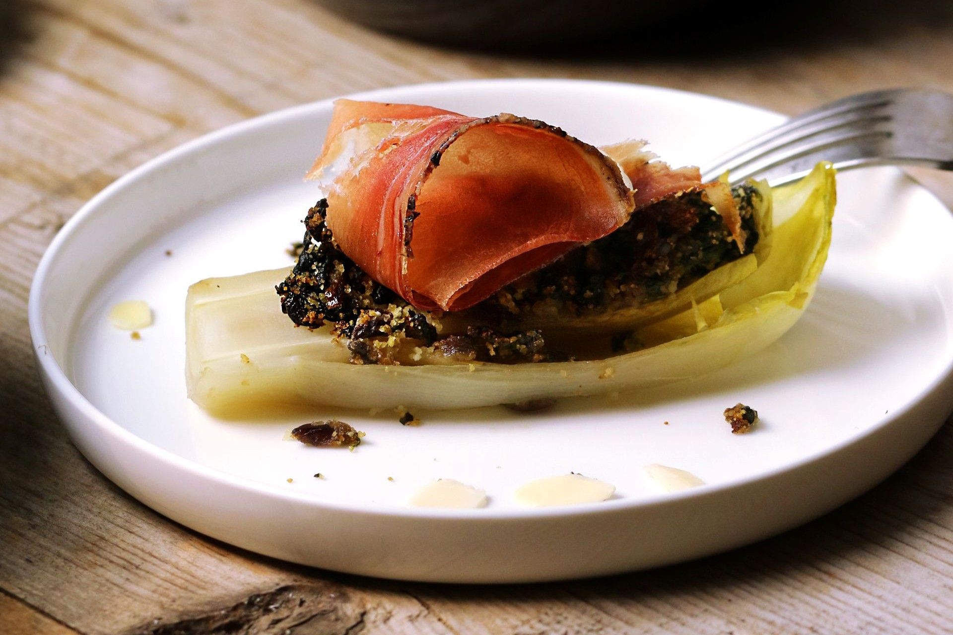 Cooked Belgian endive leaf filled with a savoury stuffing and wrapped in a slice of speck, served on a white plate.