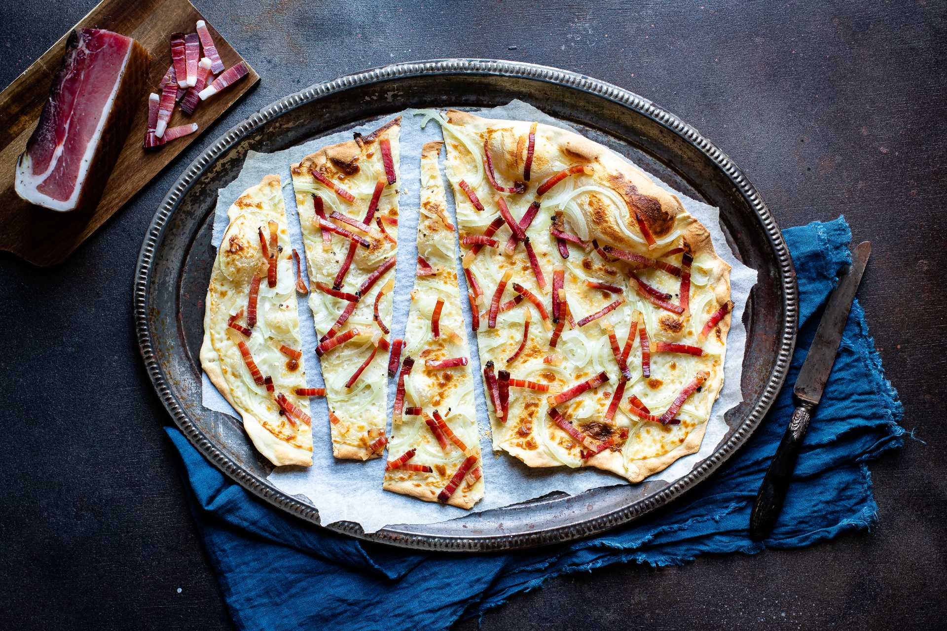 Geschnitten Flammkuchen, mit Südtiroler Speck g.g.A., goldbraunen Zwiebeln und Käsefondue.