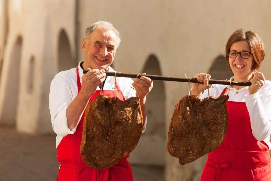 Zwei Hersteller der Mair halten Südtiroler Speck g.g.A. an Stange und lächeln im Freien