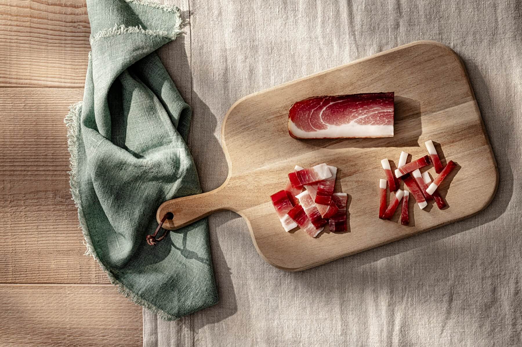 Südtiroler Speck g.g.A. in Streifen und Würfeln auf Holzbrett, daneben ein Stück Speck auf Leinentuch
