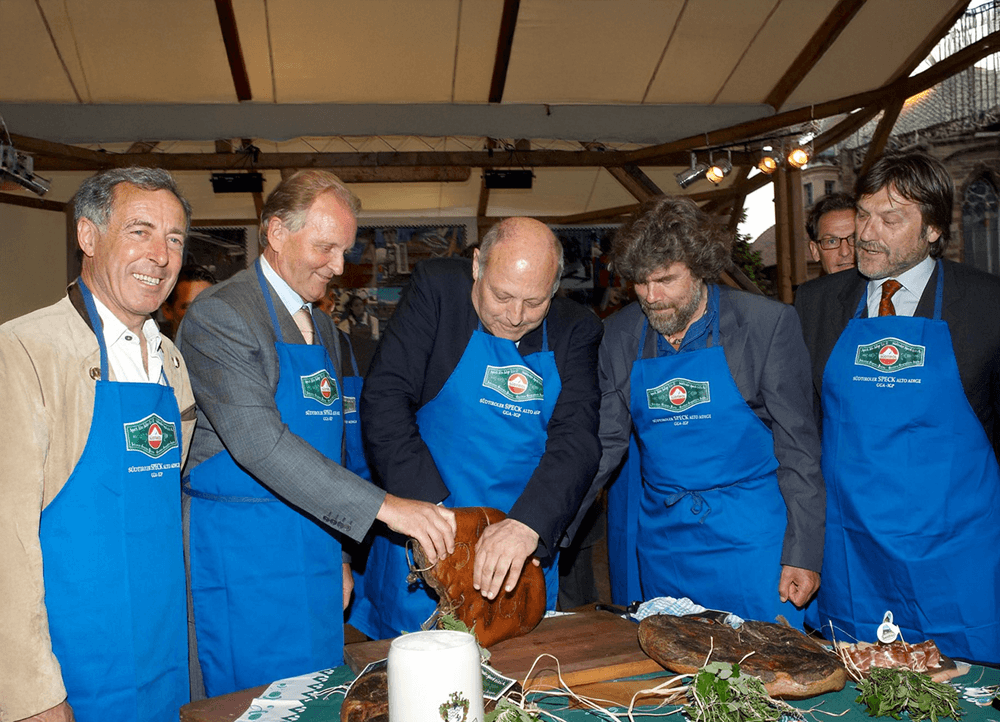Mehrere Personen mit blauen Schürzen schneiden gemeinsam ein Stück Südtiroler Speck g.g.A.