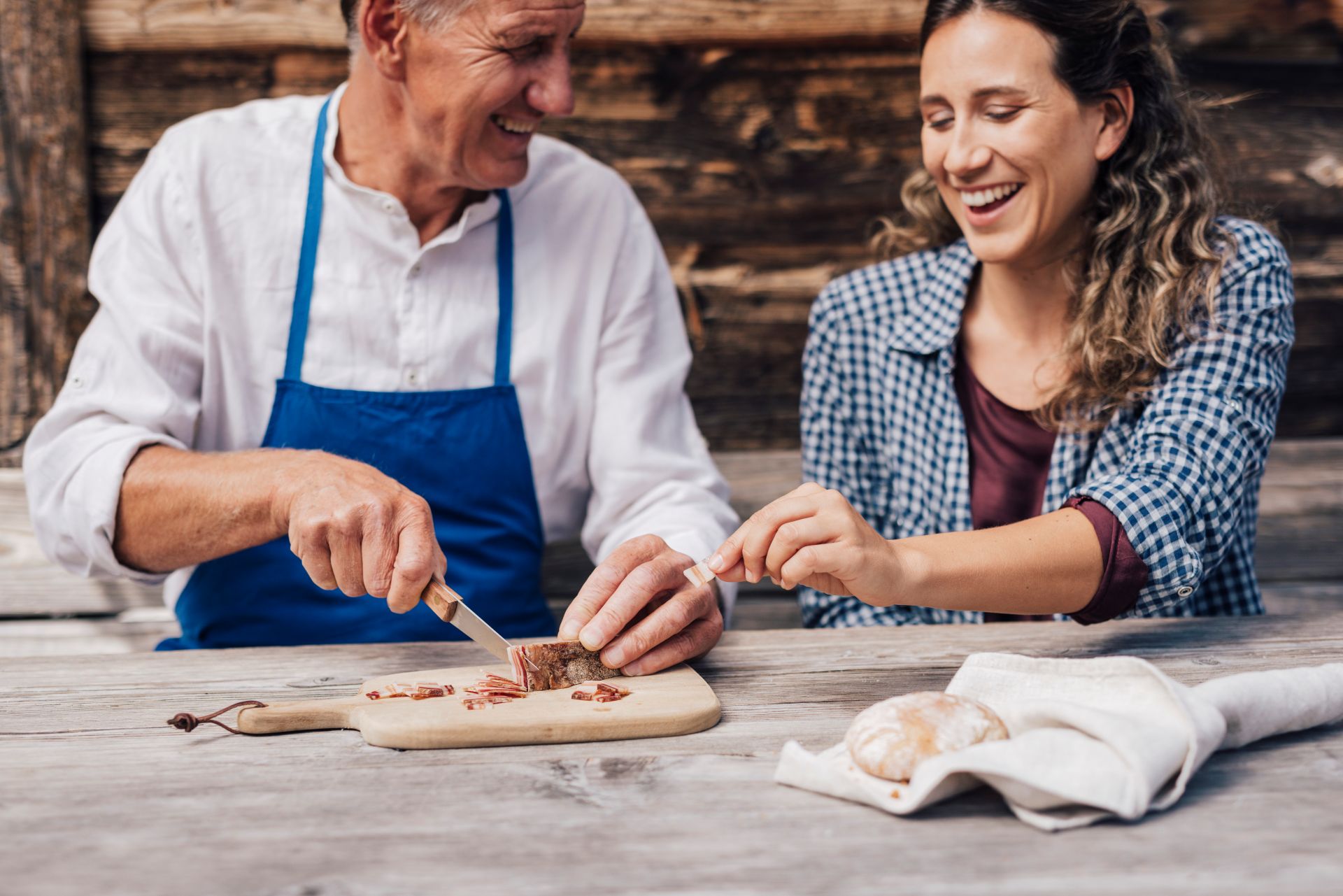 Zwei Personen lächeln am Tisch im Freien, Mann schneidet Südtiroler Speck g.g.A., Frau greift nach einer Scheibe